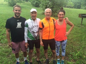 family consisting of two older men, a young man, and a young woman all stand outside on damp grass on a cloudy day wearing biking apparel. 